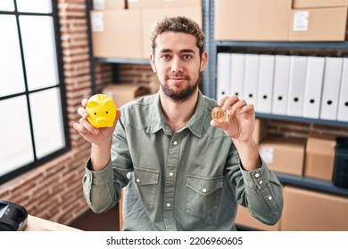 Hispanic Man With Beard Working At Small Business Ecommerce Holding Piggy Bank And Bitcoin Relaxed With Serious Expression On Face. Simple And Natural Looking At The Camera. 