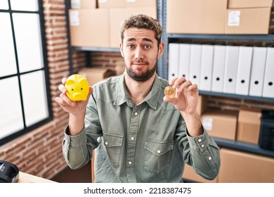 Hispanic Man With Beard Working At Small Business Ecommerce Holding Piggy Bank And Bitcoin Clueless And Confused Expression. Doubt Concept. 