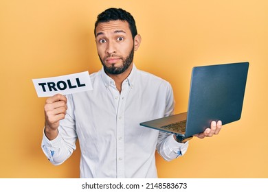 Hispanic Man With Beard Working Using Computer Laptop Holding Banner With Troll Word Clueless And Confused Expression. Doubt Concept. 