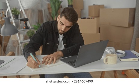 Hispanic man with beard writing in a notebook beside laptop and mug, hand on paper while leaning over boxes and ladder in a building; concentration productivity.
