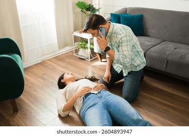 Hispanic man calling emergency while kneeling next to an unconscious woman lying on the floor after suffering from a sudden health problem