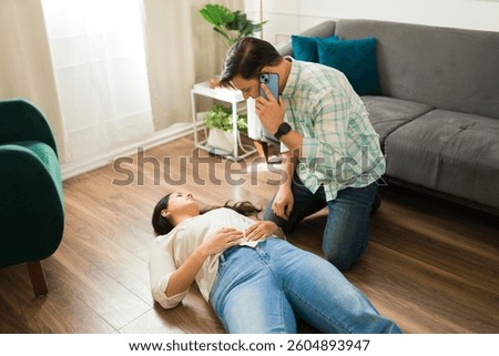Hispanic man calling emergency while kneeling next to an unconscious woman lying on the floor after suffering from a sudden health problem