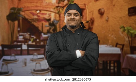 Hispanic man chef in black double breasted uniform and cap, beard visible, standing with arms crossed and downcast glance in a dining building; pride determination.