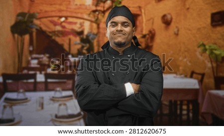 Hispanic man chef in black double breasted uniform and cap, beard visible, standing with arms crossed and downcast glance in a dining building; pride determination.