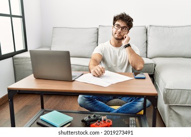 Hispanic Man Doing Papers At Home With Hand On Chin Thinking About Question, Pensive Expression. Smiling With Thoughtful Face. Doubt Concept. 