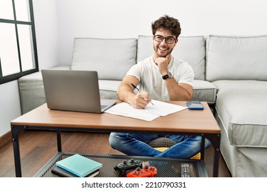 Hispanic Man Doing Papers At Home With Hand On Chin Thinking About Question, Pensive Expression. Smiling And Thoughtful Face. Doubt Concept. 