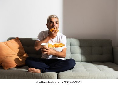 Hispanic man eating candy on a couch while whatching television. Unhealthy diet and bad habits concept. - Shutterstock ID 2052653543