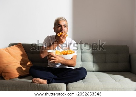 Hispanic man eating candy on a couch while whatching television. Unhealthy diet and bad habits concept.