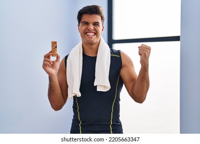 Hispanic Man Eating Protein Bar As Healthy Energy Snack Screaming Proud, Celebrating Victory And Success Very Excited With Raised Arms 