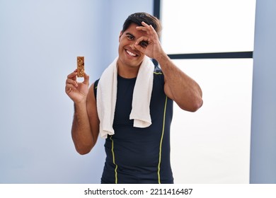 Hispanic Man Eating Protein Bar As Healthy Energy Snack Smiling Happy Doing Ok Sign With Hand On Eye Looking Through Fingers 