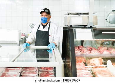 Hispanic Man With A Face Mask Standing Behind The Counter At The Butcher Shop. Male Worker Selling Quality Raw Meat At The Supermarket 