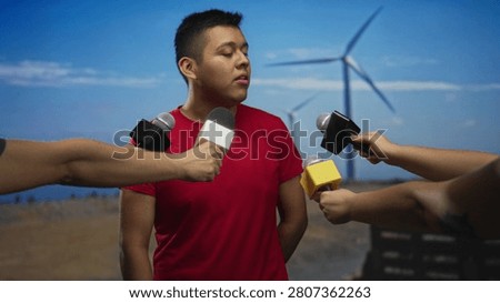 Hispanic man facing multiple microphones in a windmill field near a building; media scrutiny anxiety.