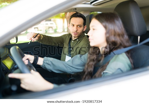 Hispanic man giving instructions and directions to a\
teen girl during her driving lessons. Male instructor talking to an\
adolescent inside a\
car