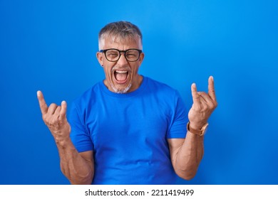 Hispanic Man With Grey Hair Standing Over Blue Background Shouting Doing Rock Symbol With Hands Up. Music Star. Heavy Music Concept. 