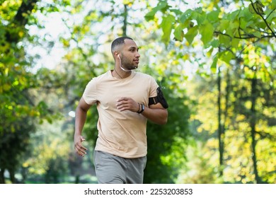 Hispanic man jogging in the park on a sunny day, runner listening to music in wired headphones, audio books and podcasts, sportsman happy with an active weekend. - Powered by Shutterstock