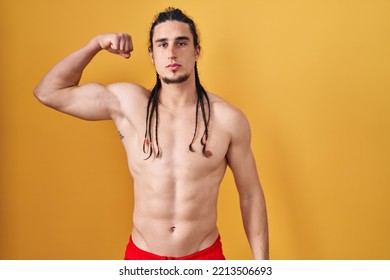 Hispanic Man With Long Hair Standing Shirtless Over Yellow Background Strong Person Showing Arm Muscle, Confident And Proud Of Power 