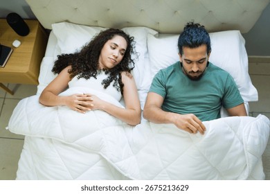Hispanic man lying in bed, grappling with erectile dysfunction, while his concerned wife sits beside him, sharing the weight of their worries