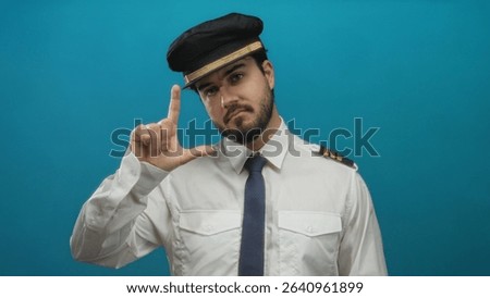 Hispanic man in a pilot uniform makes a l gesture toward the camera against a blue background, showcasing humor and authority with a playful expression.