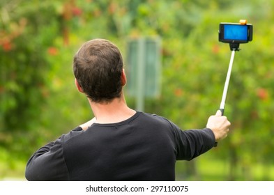 Hispanic Man Posing With Selfie Stick In Park Environment, His Back Facing Camera.