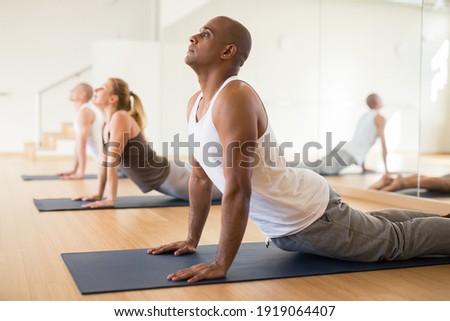Hispanic man practicing yoga postures during group training at gym, performing stretching asana Urdhva Mukha Shvanasana (Upward Facing Dog Pose)