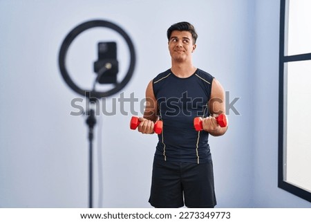 Hispanic man recording training tutorial at the gym smiling looking to the side and staring away thinking. 