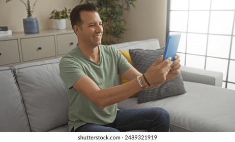 Hispanic man sitting indoors in a modern living room, holding a tablet and smiling. - Powered by Shutterstock