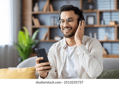 Hispanic man sitting on couch wearing headphones, using smartphone. He is smiling, appearing relaxed and happy, enjoying music indoors. Scene conveys leisure, technology, and modern lifestyle. - Powered by Shutterstock