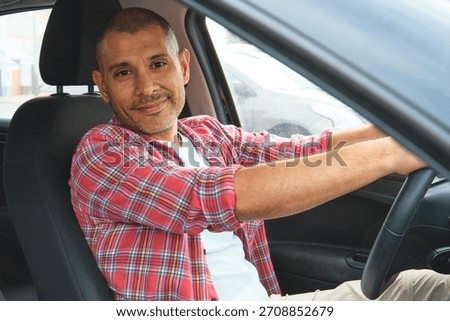 Hispanic man smiling while driving a car, wearing a red plaid shirt
