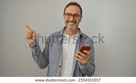Hispanic man smiling while holding a smartphone and pointing with a finger over isolated white background
