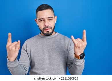 Hispanic Man Standing Over Blue Background Shouting With Crazy Expression Doing Rock Symbol With Hands Up. Music Star. Heavy Music Concept. 