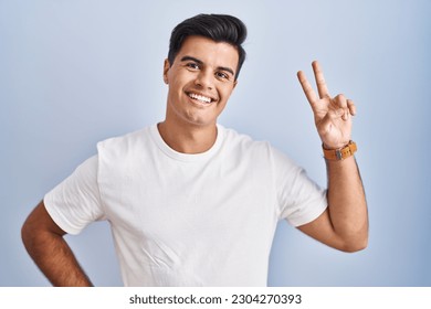 Hispanic man standing over blue background smiling looking to the camera showing fingers doing victory sign. number two. 