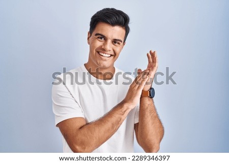 Hispanic man standing over blue background clapping and applauding happy and joyful, smiling proud hands together 