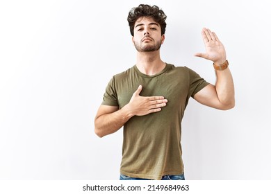 Hispanic man standing over isolated white background swearing with hand on chest and open palm, making a loyalty promise oath 