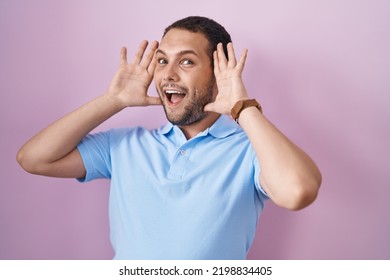 Hispanic Man Standing Over Pink Background Smiling Cheerful Playing Peek A Boo With Hands Showing Face. Surprised And Exited 