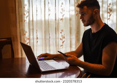 Hispanic man studying using multiscreen with laptop and smartphone close to the window