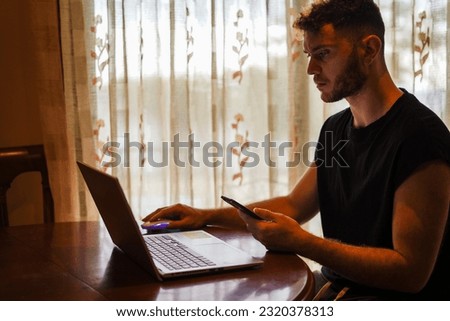 Hispanic man studying using multiscreen with laptop and smartphone close to the window