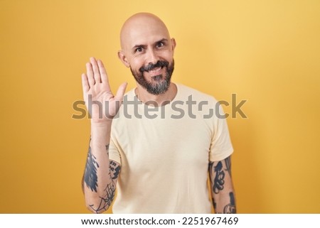 Hispanic man with tattoos standing over yellow background waiving saying hello happy and smiling, friendly welcome gesture 
