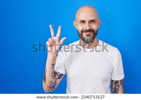 Hispanic man with tattoos standing over blue background showing and pointing up with fingers number three while smiling confident and happy. 