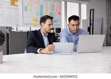 Hispanic man using digital tablet while his Asian colleague working on laptop, looking focused in their joint tasks, engaged in collaborative work using two modern devices in office. Digital workflow