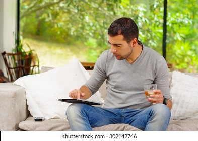 Hispanic Man Wearing Denim Jeans And Grey Sweater Sitting In Sofa With Tablet Resting On Knee Looking At Screen Holding Glass Of Dark Colored Beverage.