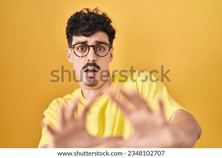 Hispanic man wearing glasses standing over yellow background afraid and terrified with fear expression stop gesture with hands, shouting in shock. panic concept. 