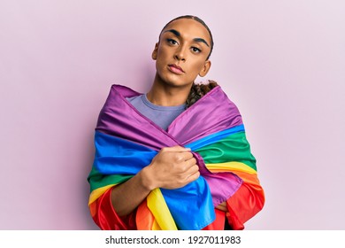 Hispanic Man Wearing Make Up And Long Hair Wrapped In Rainbow Lgbtq Flag Relaxed With Serious Expression On Face. Simple And Natural Looking At The Camera. 