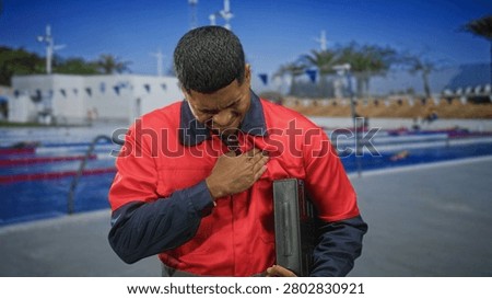 Hispanic man worker in red uniform clutching chest for heartattack while holding toolbox at outdoor pool building; distress concern.