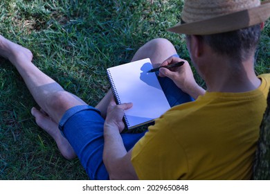 Hispanic Mature Adult Man With A Hat Sitting On A Park Lawn And Leaning On A Tree With A Blank Notebook In One Hand And A Pen In The Other.