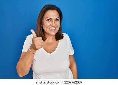 Mujer hispana madura parada sobre fondo azul haciendo gesto de pulgares hacia arriba con la mano. expresión de aprobación mirando a la cámara mostrando éxito. 