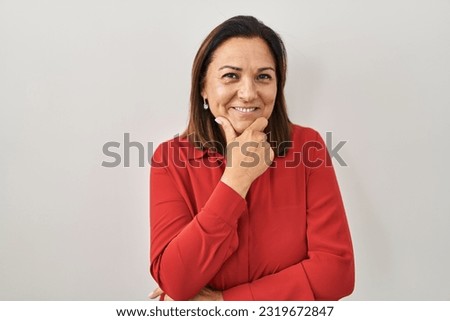 Hispanic mature woman standing over white background looking confident at the camera smiling with crossed arms and hand raised on chin. thinking positive. 