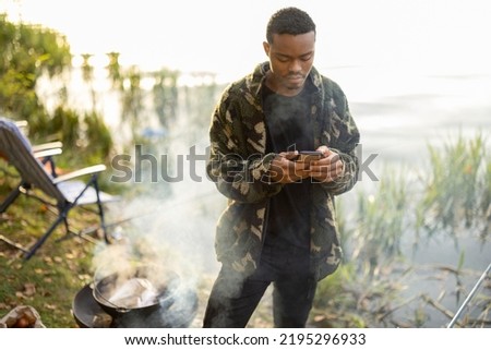 Hispanic men using smartphones during resting on river or pond coast. Men fishing and cooking on river coast at autumn. Leisure and weekend in nature