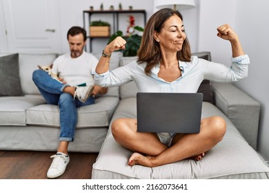 Hispanic Middle Age Couple At Home, Woman Using Laptop Showing Arms Muscles Smiling Proud. Fitness Concept. 