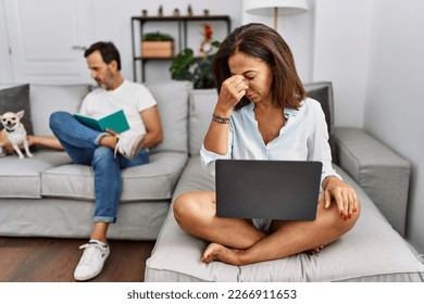 Pareja hispánica de mediana edad en casa, mujer usando laptop cansada frotando nariz y ojos sintiendo fatiga y dolor de cabeza. concepto de estrés y frustración. 