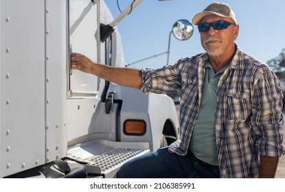 Hispanic Middle Aged Semi Truck Driver Sits Next To Truck. Experienced Senior Trucker Next To Big Rig At The Job. Portrait Of Transportation And Logistics Worker Next To Equipment On A Sunny Day.